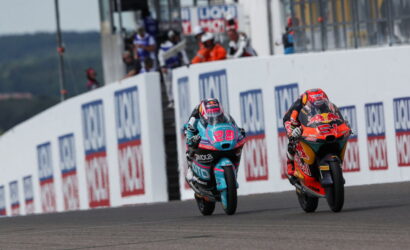 Two motorcycle racers sprint side by side on a race track, leaning into the turn past sponsor boards.