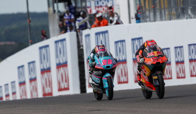 Two motorcycle racers sprint side by side on a race track, leaning into the turn past sponsor boards.