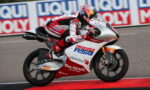 Motorcycle racer on a red-and-white Honda bike leaning into a turn on a race track, sponsor logos visible along the bodywork and rider’s suit.