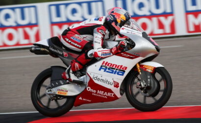 Motorcycle racer on a red-and-white Honda bike leaning into a turn on a race track, sponsor logos visible along the bodywork and rider’s suit.
