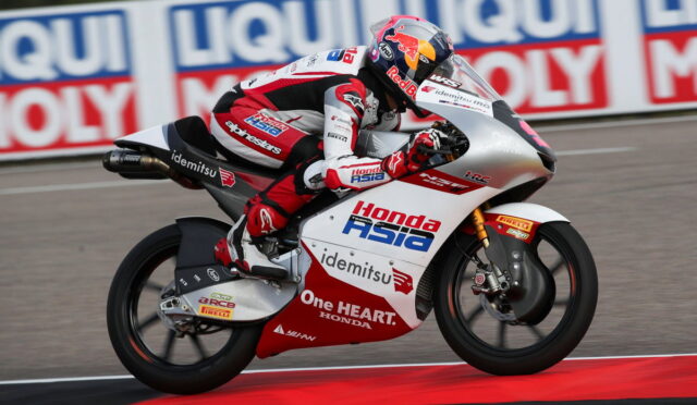 Motorcycle racer on a red-and-white Honda bike leaning into a turn on a race track, sponsor logos visible along the bodywork and rider’s suit.