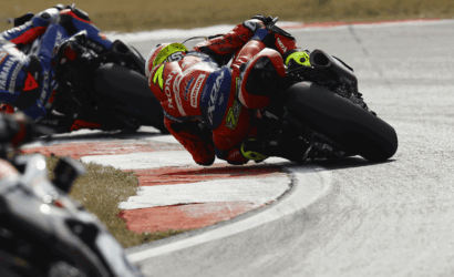Motorcycle racers lean into a sharp turn on a race track, centered on a red‑and‑white bike with a neon‑green helmet in a tight corner.