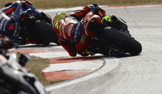 Motorcycle racers lean into a sharp turn on a race track, centered on a red‑and‑white bike with a neon‑green helmet in a tight corner.