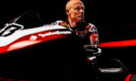 Professional motorcycle racer in a racing suit sits beside a red-and-white race bike against a dark background, gazing to the right.