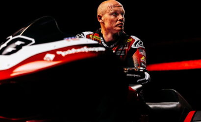 Professional motorcycle racer in a racing suit sits beside a red-and-white race bike against a dark background, gazing to the right.
