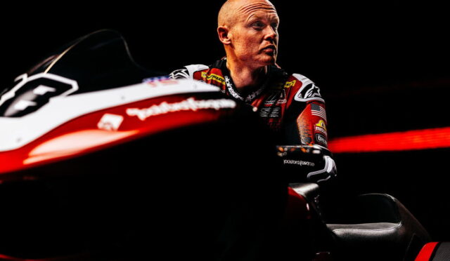 Professional motorcycle racer in a racing suit sits beside a red-and-white race bike against a dark background, gazing to the right.