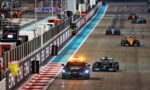 Formula 1 cars race along a night-lit track; a safety car with flashing orange lights leads the pack past barriers and sponsor boards (Etihad).