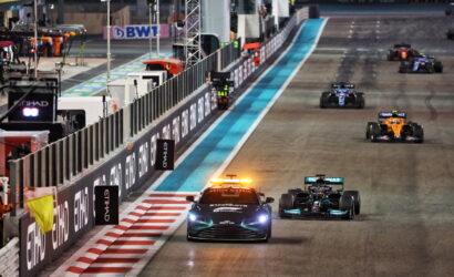 Formula 1 cars race along a night-lit track; a safety car with flashing orange lights leads the pack past barriers and sponsor boards (Etihad).