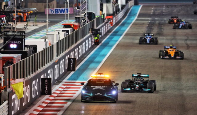 Formula 1 cars race along a night-lit track; a safety car with flashing orange lights leads the pack past barriers and sponsor boards (Etihad).