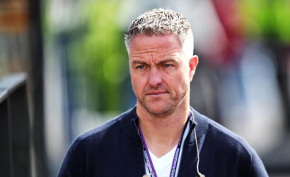 Casual portrait of a man with short gray hair and stubble, wearing a navy sweater and a lanyard with earphones outdoors.