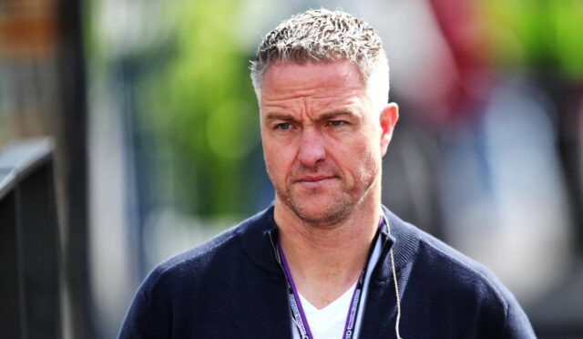 Casual portrait of a man with short gray hair and stubble, wearing a navy sweater and a lanyard with earphones outdoors.