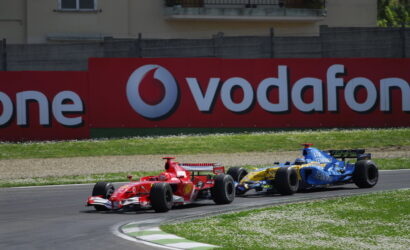 Red Formula 1 car leading a blue Renault behind on a race track with a large Vodafone sign in the background
