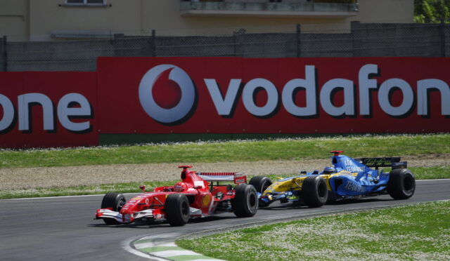 Red Formula 1 car leading a blue Renault behind on a race track with a large Vodafone sign in the background