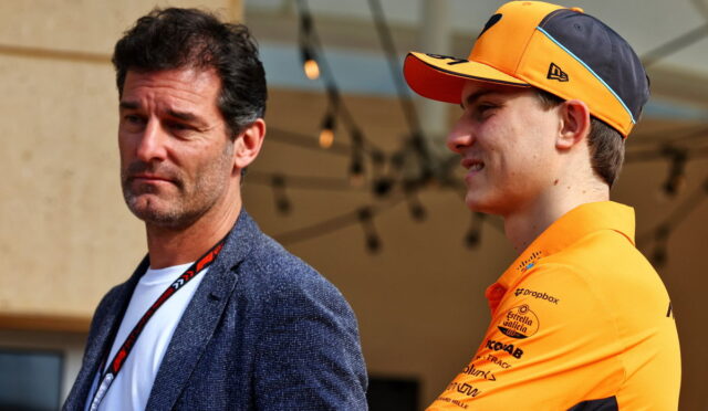 Man in a blue blazer and white shirt stands beside a young cyclist in an orange cap and jersey at an outdoor event.