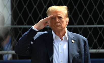Man in a dark suit saluting with his right hand, standing before a chain-link fence.
