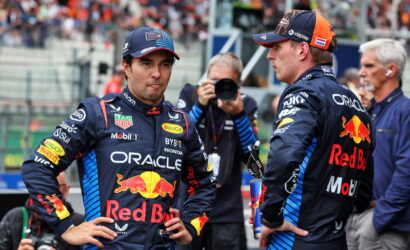 Two Red Bull Racing drivers in navy suits with Red Bull logos stand facing each other on a pit lane while a photographer shoots in the background.