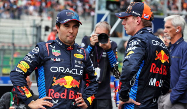 Two Red Bull Racing drivers in navy suits with Red Bull logos stand facing each other on a pit lane while a photographer shoots in the background.