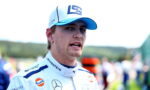 Race car driver in a white racing suit with sponsor logos and a blue cap, speaking or gesturing outdoors in a racing paddock.
