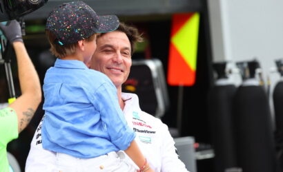 Smiling man in a white team polo holds a young boy in a blue shirt and a colorful confetti-pattern cap in a race team pit area.