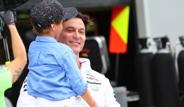 Smiling man in a white team polo holds a young boy in a blue shirt and a colorful confetti-pattern cap in a race team pit area.