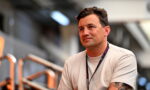 Man with short dark hair and a sleeves tattoo, wearing a beige t‑shirt and a lanyard, seated in a workshop or backstage area.
