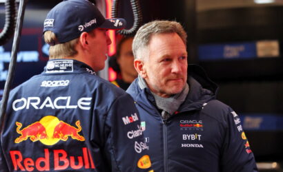 Two Red Bull Racing crew members in branded jackets chat in the pits, Oracle and Red Bull logos visible.