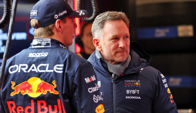 Two Red Bull Racing crew members in branded jackets chat in the pits, Oracle and Red Bull logos visible.