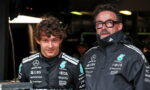 Two race team members in black Mercedes jackets pose in a dim garage beside a monitor and equipment, facing the camera.