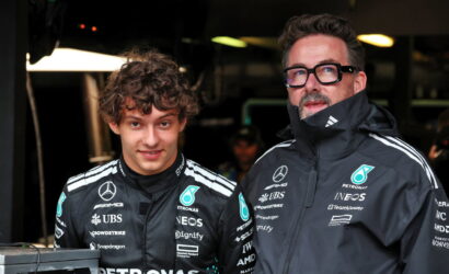 Two race team members in black Mercedes jackets pose in a dim garage beside a monitor and equipment, facing the camera.