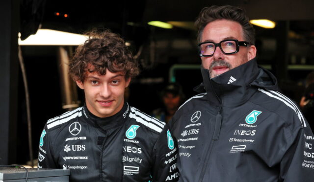 Two race team members in black Mercedes jackets pose in a dim garage beside a monitor and equipment, facing the camera.