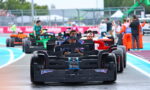 Front black Formula-style race car leads a line of cars on a wet pit-lane, drivers visible in cockpits. Progressing cars in various colors follow behind, with crew and spectators in the background.