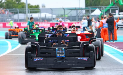 Front black Formula-style race car leads a line of cars on a wet pit-lane, drivers visible in cockpits. Progressing cars in various colors follow behind, with crew and spectators in the background.