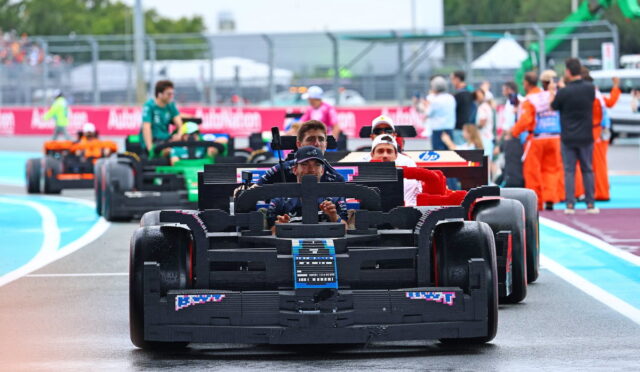 Front black Formula-style race car leads a line of cars on a wet pit-lane, drivers visible in cockpits. Progressing cars in various colors follow behind, with crew and spectators in the background.