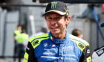 Male race car driver in a blue and neon-yellow racing suit, wearing a Monster Energy cap, smiling on a pit lane.