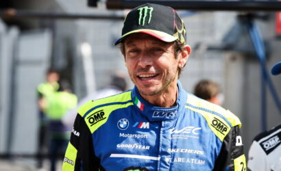 Male race car driver in a blue and neon-yellow racing suit, wearing a Monster Energy cap, smiling on a pit lane.