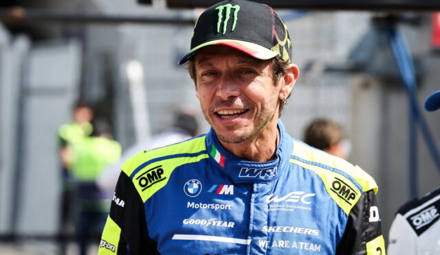 Male race car driver in a blue and neon-yellow racing suit, wearing a Monster Energy cap, smiling on a pit lane.