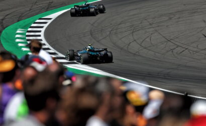 Two blue Formula 1 cars race around a tight curve on a track, with a blurred crowd in the foreground and tire marks on the asphalt.