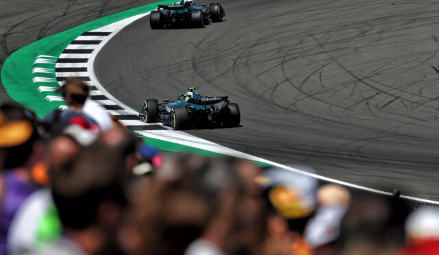 Two blue Formula 1 cars race around a tight curve on a track, with a blurred crowd in the foreground and tire marks on the asphalt.