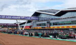Pit lane at a race circuit with teams and crew lined up, large 'LAS VEGAS' banners overhead and a modern grandstand facade in the background.