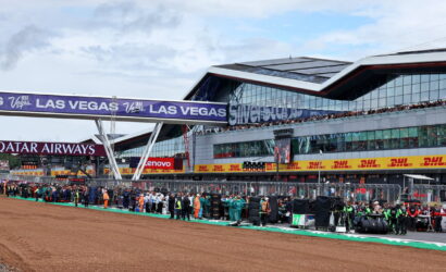 Pit lane at a race circuit with teams and crew lined up, large 'LAS VEGAS' banners overhead and a modern grandstand facade in the background.