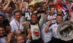 Celebrating race team after win: driver in white suit raises a gold trophy above his head, flanked by cheering teammates and fans, with a silver decorative tray visible on the right.