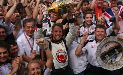 Celebrating race team after win: driver in white suit raises a gold trophy above his head, flanked by cheering teammates and fans, with a silver decorative tray visible on the right.