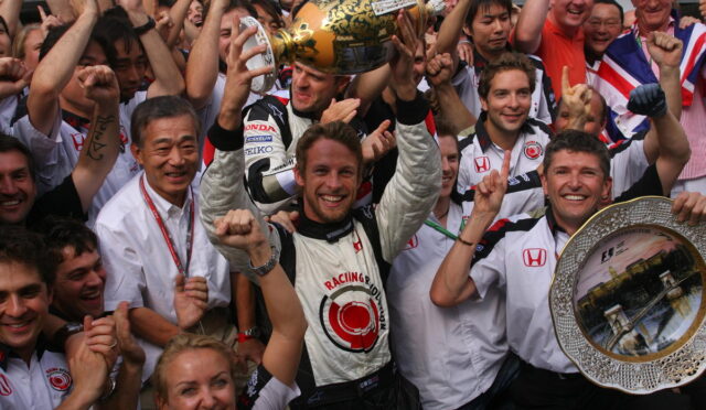 Celebrating race team after win: driver in white suit raises a gold trophy above his head, flanked by cheering teammates and fans, with a silver decorative tray visible on the right.