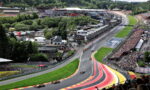 Aerial view of a Formula 1 race on a winding track with colorful red, yellow, and pink run-off stripes, packed grandstands, and pit buildings in the background.