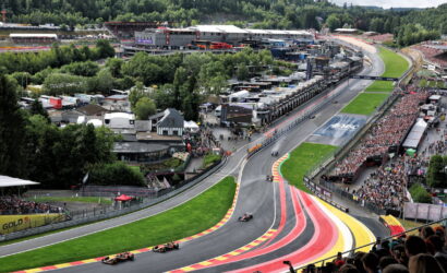 Aerial view of a Formula 1 race on a winding track with colorful red, yellow, and pink run-off stripes, packed grandstands, and pit buildings in the background.