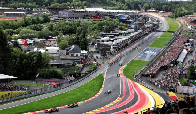 Aerial view of a Formula 1 race on a winding track with colorful red, yellow, and pink run-off stripes, packed grandstands, and pit buildings in the background.