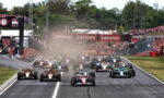 Formula 1 cars line up and start a race on a runway-like track as dust rises, with Lenovo banners and cheering crowds in the background.