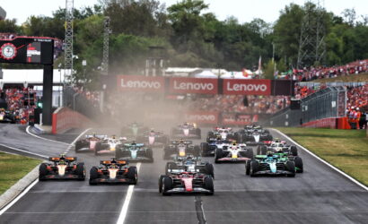 Formula 1 cars line up and start a race on a runway-like track as dust rises, with Lenovo banners and cheering crowds in the background.