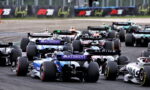 F1 race cars tightly packed at the start on a dark tarmac circuit, sponsor logos visible on rear wings, creating a chaotic grid moment.