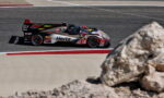 Prototype race car (Cadillac DPi) with Hertz sponsor speeding around a turn on an asphalt track, rock foreground on the right side.